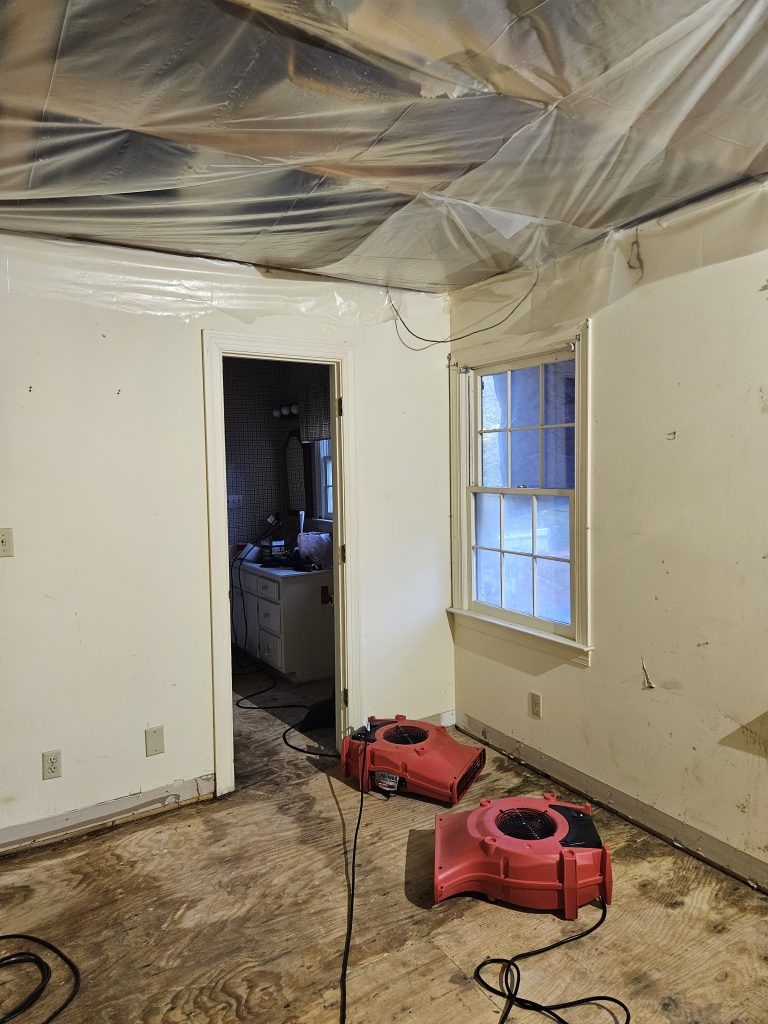 A partially remodeled room with bare wooden floors, featuring two industrial red fans for drying. Plastic sheeting covers the ceiling, indicating ongoing repairs.