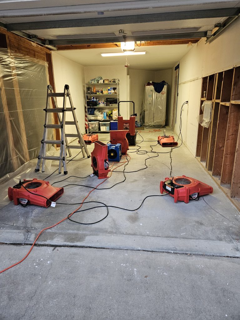 A garage equipped with multiple red air movers and a dehumidifier, surrounded by tools and supplies, indicating ongoing water damage restoration work.