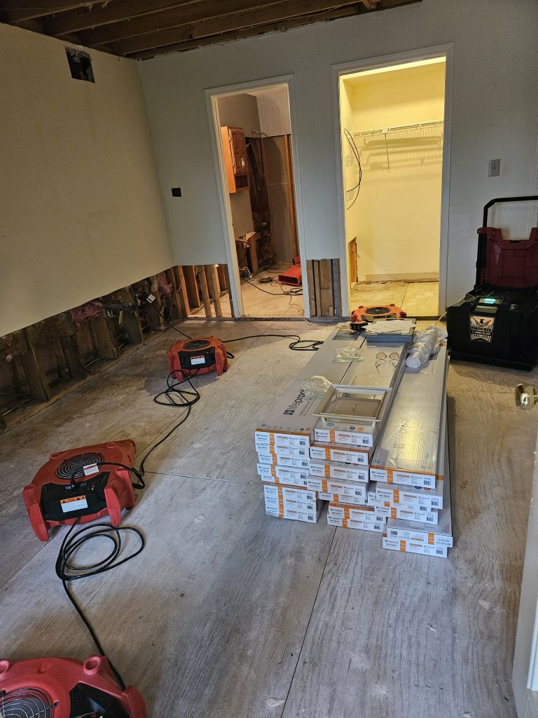 Renovation scene showing an unfinished room with exposed walls, construction equipment, and stacked flooring materials ready for installation.