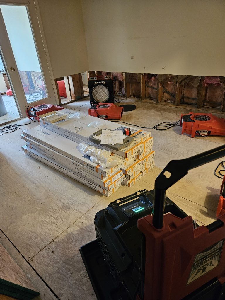 Renovation in progress: stacked flooring materials await installation, with fans drying the space. Visible damage to the wall indicates prior water issues.