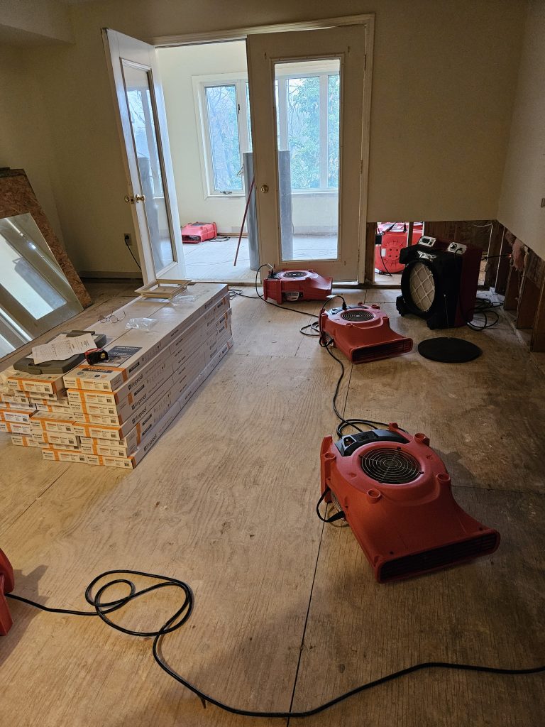 Surfaces with wood flooring and construction supplies; red air movers are set up for drying in a remodeling space with large windows.