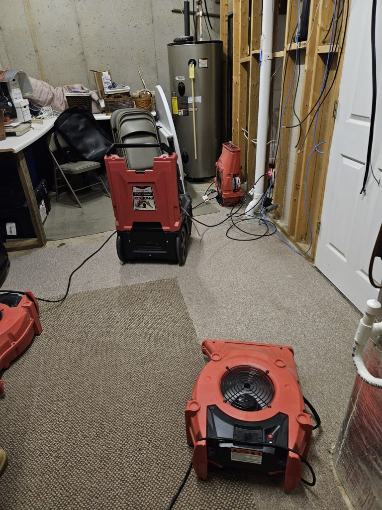 Water damage restoration equipment is set up in a basement. Two red air movers are positioned on the carpet near a water heater, indicating ongoing repairs.