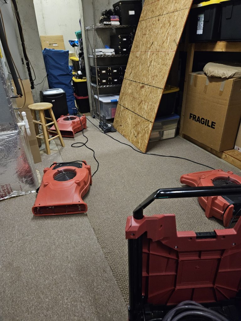 Red air circulation fans and a vacuum sit on carpet in a cluttered basement space, indicating cleanup or drying work in progress.