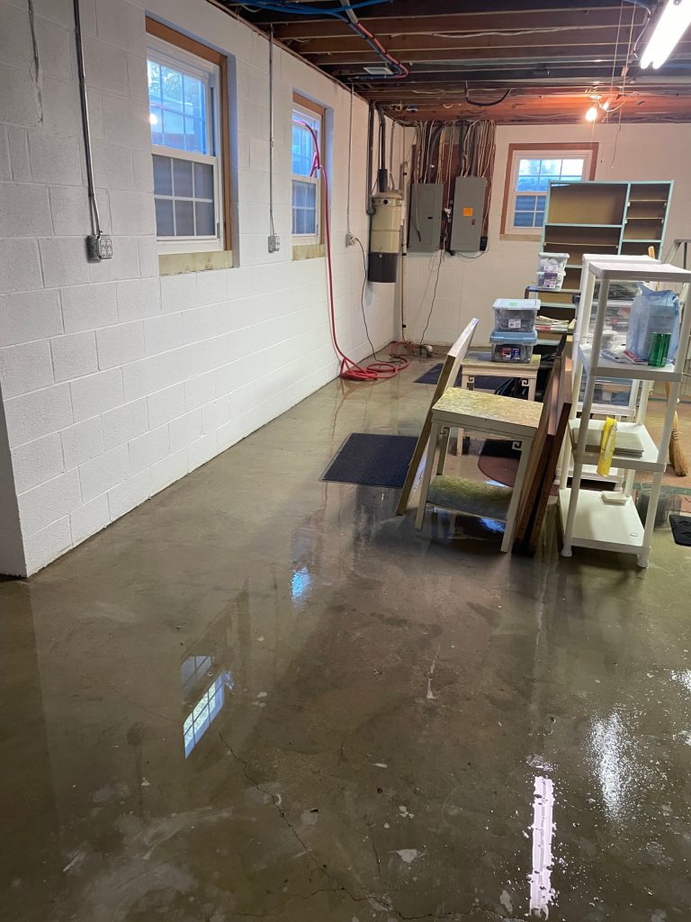 Water covers the concrete floor of a basement, reflecting light from windows above. Shelves and tools are visible, indicating a workspace.