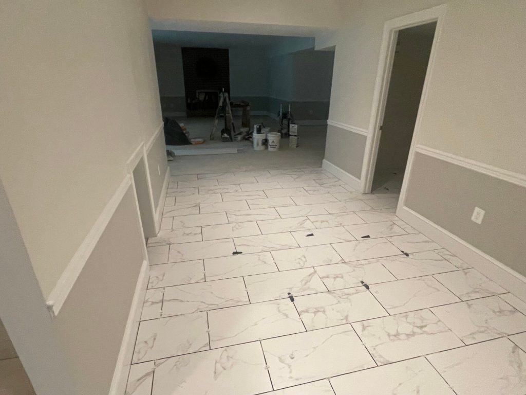 Renovated room with newly laid white marble-patterned tile flooring. Wall trim is partially visible, with tools and paint supplies in the background.