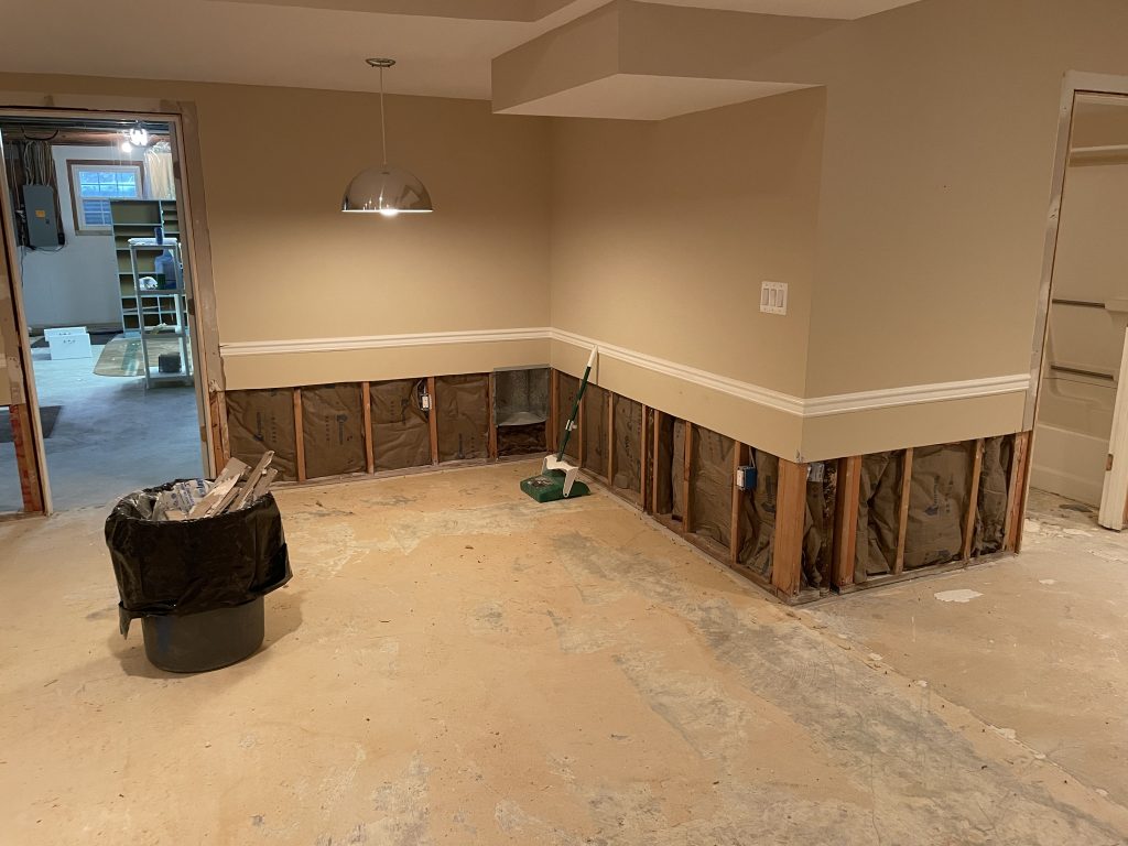 Basement under renovation, featuring exposed walls and a bare floor. A trash can and broom are present, indicating construction activity.
