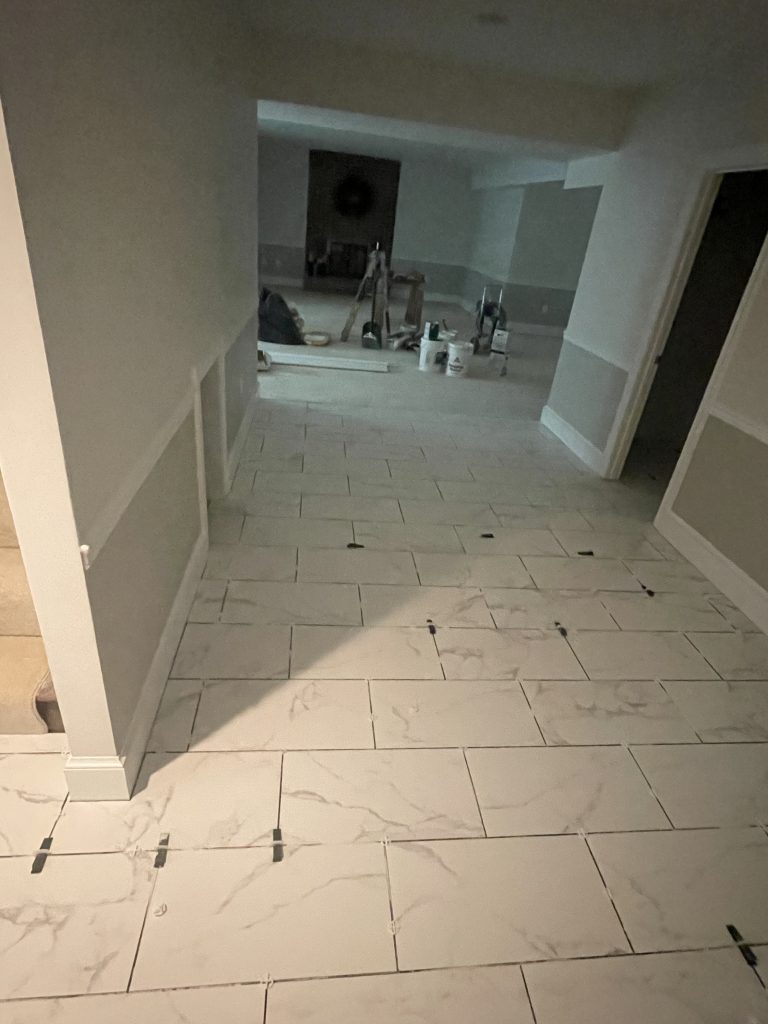 Light-colored marble tiles are laid on the floor of a hallway, with spacers in place. A partially furnished room is visible in the background, indicating renovation.