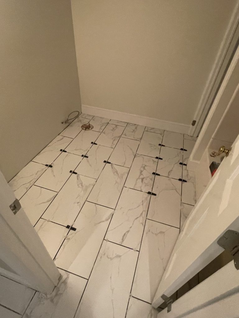 Newly tiled flooring in a small room features white marble-patterned tiles with black spacers. A plumbing fixture is visible in one corner.