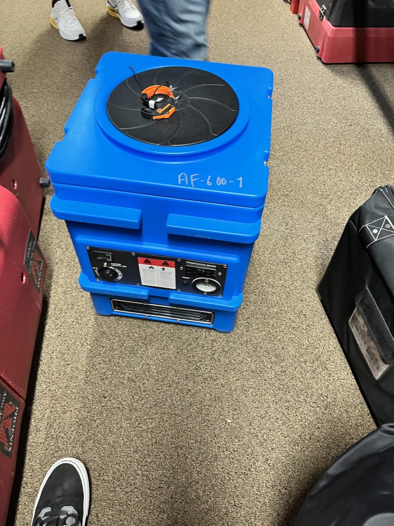 Blue equipment case with knobs and controls, topped with a black fan cover. Positioned on a carpeted floor amid other cases, it suggests functionality.