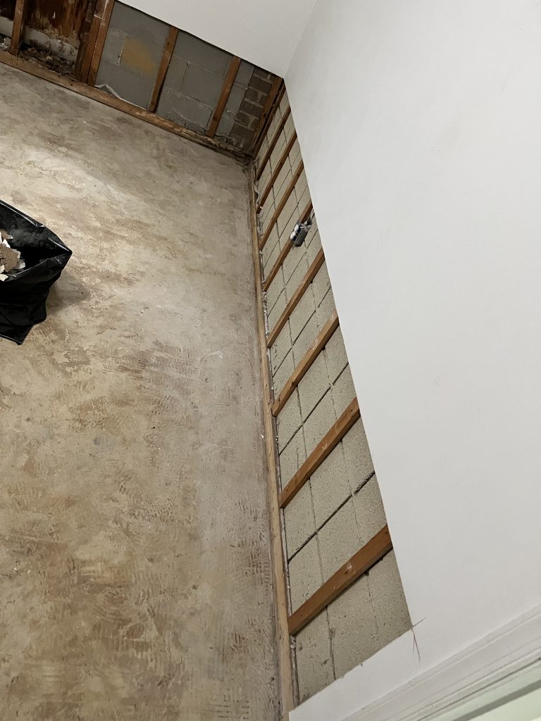 Renovated indoor space showing bare concrete floor and exposed wall studs with cinderblock. A black garbage bag holds debris.